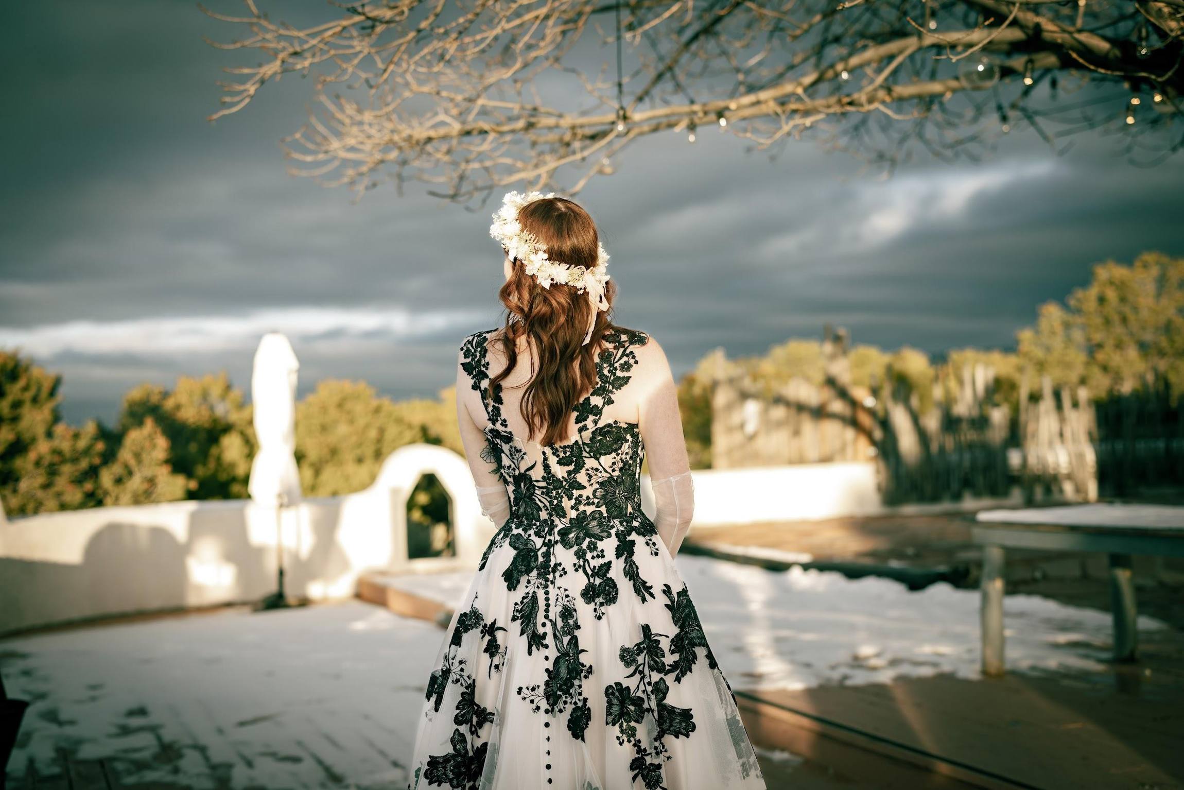 Bride in a black floral gown and flower crown faces a stormy sky