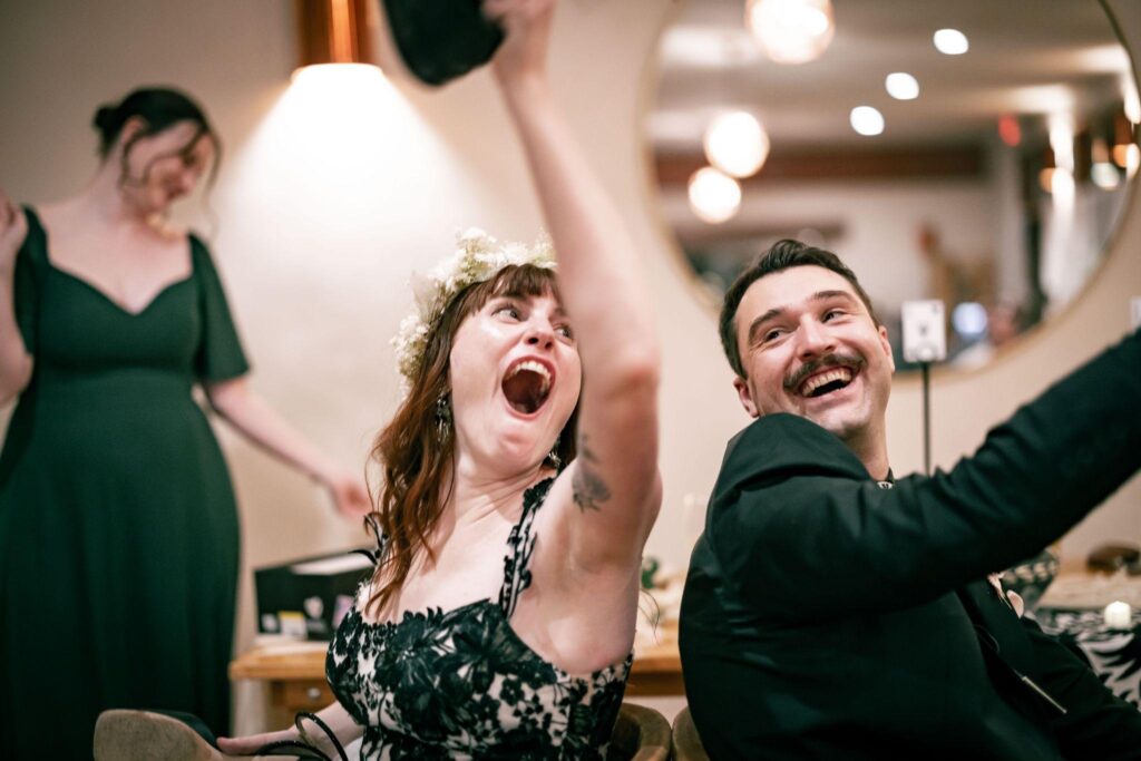 Bride in a flower crown cheers with a hat raised beside laughing groom